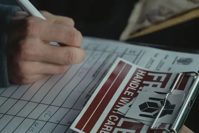 A hand signing a dispatch checklist on a clipboard, beside a sheet of fragile handling labels.