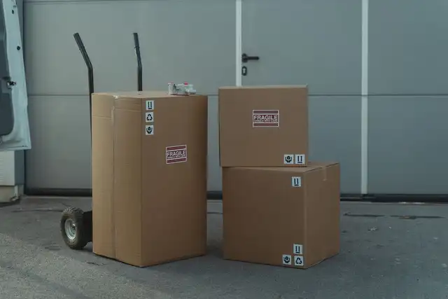 Three sealed cardboard cartons marked fragile, stacked beside a sack truck at the open rear of a van.