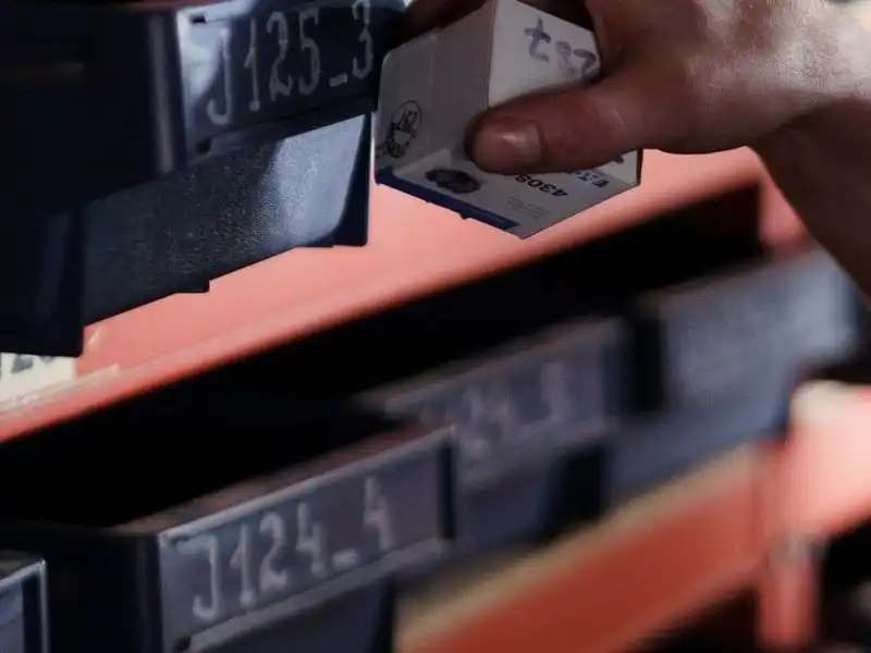 A hand lifting a small boxed component out of bin J125_3 on warehouse racking, among rows of identically labelled bins.